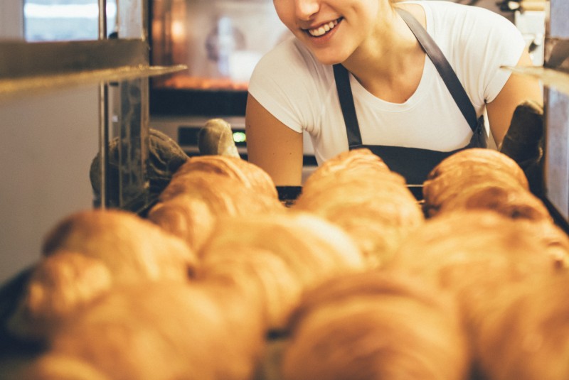 Blick aus einem Industrie-Backofen voller Croissants auf die Bäckerin, die das fertige Blech mit Handschuhen herausholt