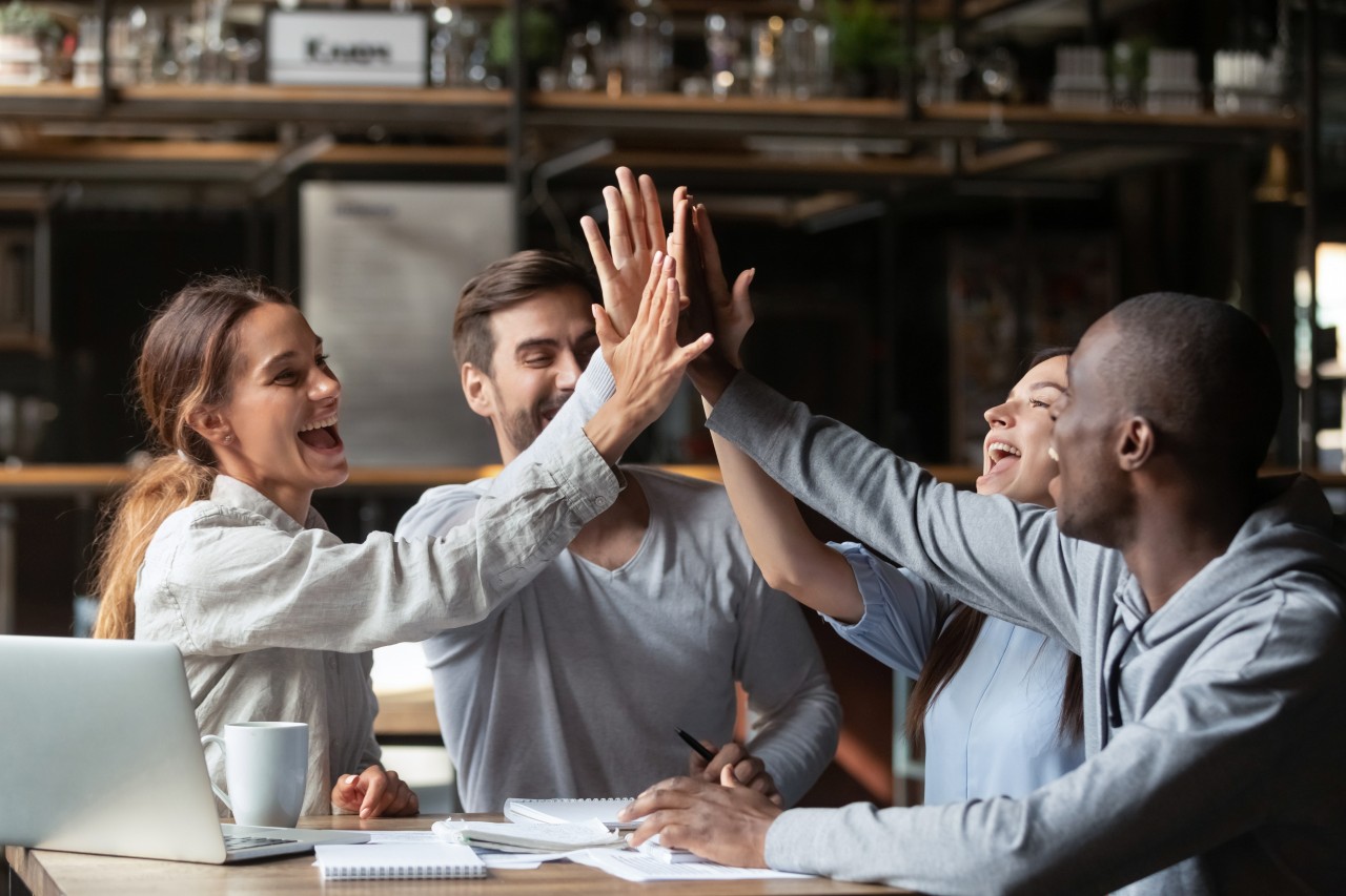 Zwei Männer und zwei Frauen sitzen mit Laptop, Notizblöcken und Kaffee gemeinsam am Tisch und geben sich ein High-Five