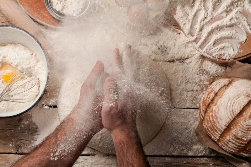 Bemehlte Unterarme und Hände eines Menschen über einem Teig, daneben weitere Backzutaten, Utensilien und ein Laib Brot